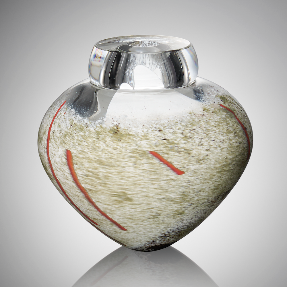 A thick clear hand blown glass vessel featuring subtle shades of gray and white glass with streaks of red glass cane stands against a light gray background.