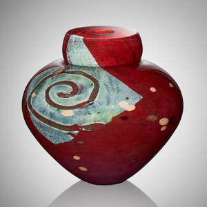 A deep red blown glass vessel features a square of silver leaf decorated with a swirl of clear glass, revealing the red coloration beneath. The surface is adorned with pops of burgundy and apricot glasses.