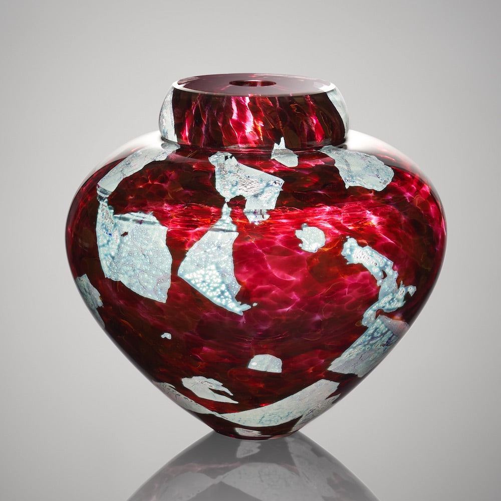 A ruby red hand blown glass vessel featuring layers of silver foil stands against a light gray background.