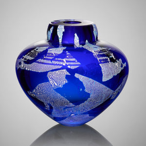 A vibrant blue hand blown glass vessel featuring layers of silver leaf stands against a gray background.
