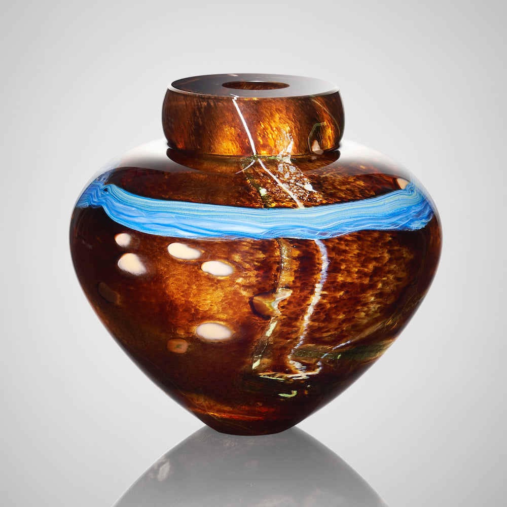 An earthy brown hand blown glass vessel featuring a band of silver blue glass stands against a gray background.