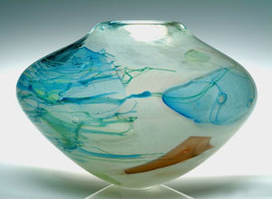 A large blue and white blown glass vessel with handpulled blue glass canes and shards of blues and browns stands against a light gray background.