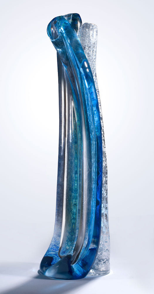 A tall hand blown glass sculpture featuring layers of blue, silver, and clear glass stands against a white background.