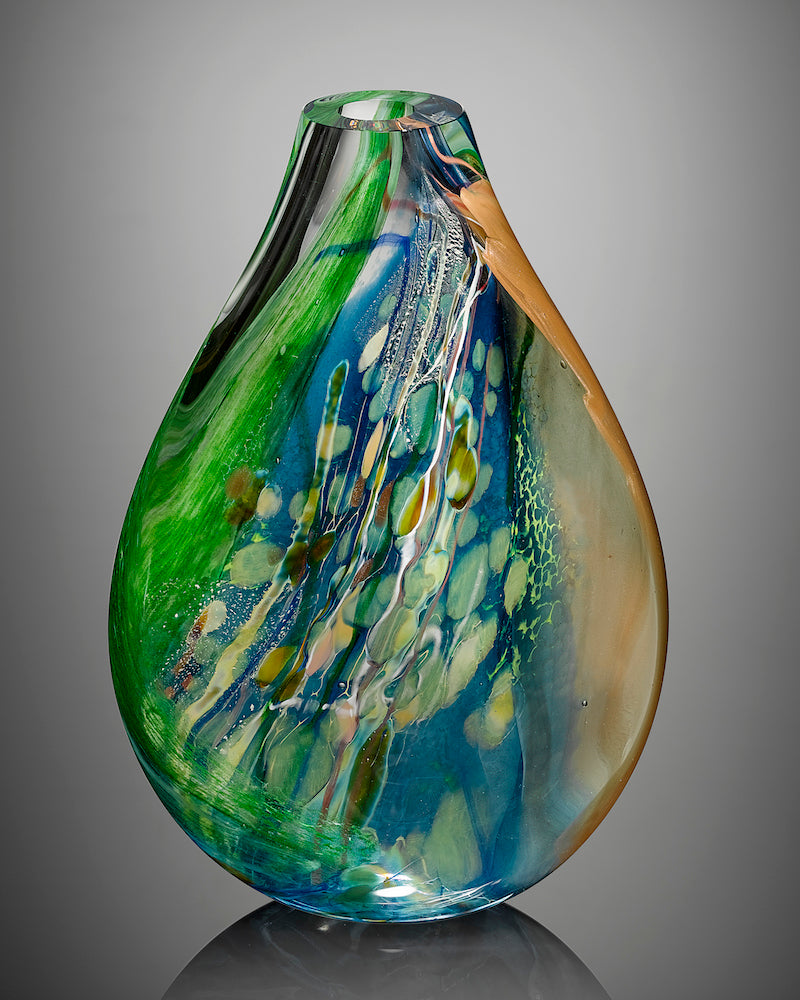 A sculptural blown glass vessel featuring blue, green, and clear glass stands against a gray background.