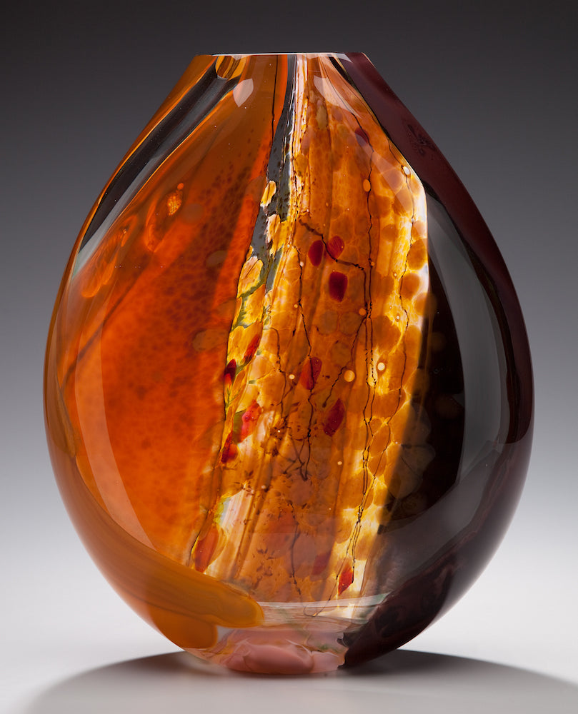 A hand blown glass vessel featuring autumn colors of orange, gold, and brown glass stands against a gray background.