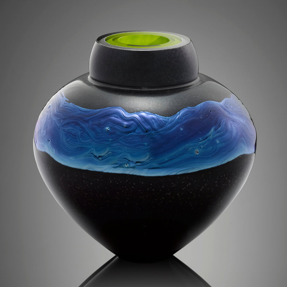 A black hand blown glass vessel features a band of silver blue glass and an interior layer of bright green glass.