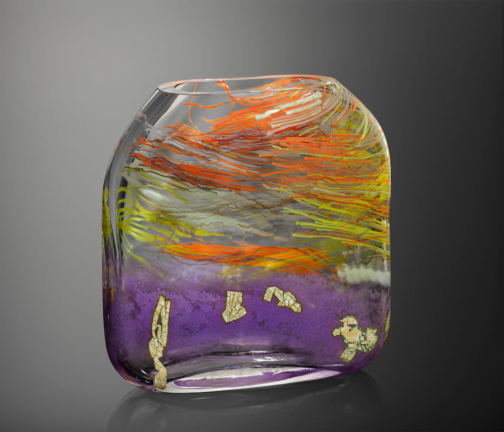 A large hand blown glass vessel molded into a square form features vibrant streaks of orange, yellow, red, and white glass cane in thick clear glass, featuring a band of orchid purple glass at the bottom with flecks of silver leaf.