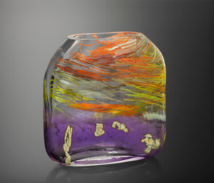 A large hand blown glass vessel molded into a square form features vibrant streaks of orange, yellow, red, and white glass cane in thick clear glass, featuring a band of orchid purple glass at the bottom with flecks of silver leaf.