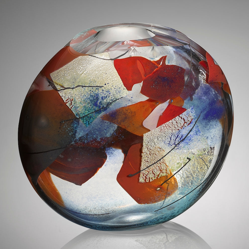 A thick clear hand blown glass vessel featuring shards of red and blue glass, layers of silver leaf, and veins of black glass cane stands against a gray background.