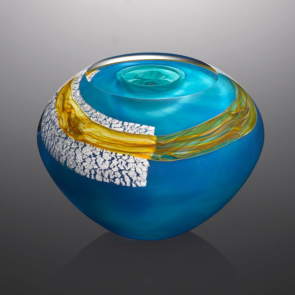 A bright blue hand blown glass vessel featuring a sheet of silver leaf and a band of golden glass stands against a gray background.