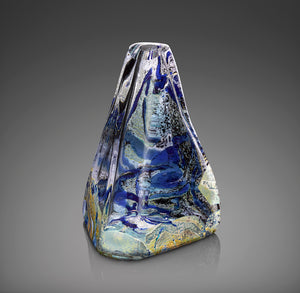 A hand blown glass vessel molded into a tall pyramid shape features layers of deep cobalt blue, shimmering silver, and white glass powder encased in thick clear glass.