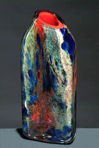 A tall rectangular blown glass vessel features layers of red, blue, white, and green glass powders with a volcanic red interior.