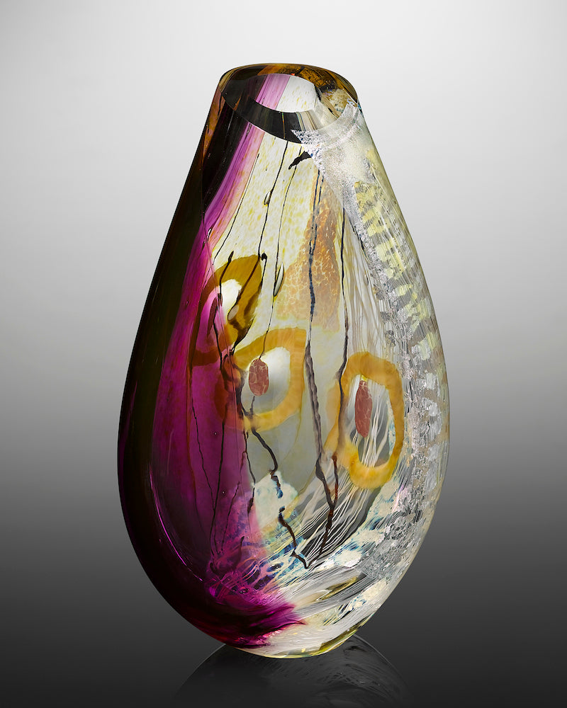 A tall blown glass vessel features translucent purple glass, delicate white glass cane, layers of silver foil, and playful line drawings in glass cane with pops of purple and gold glass.