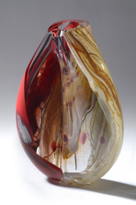 A thick sculptural blown glass vessel features deep red, soft gray, and warm brown tones with pops of red and white glasses. The form is slightly twisted, evoking the movement of a woman's hips.