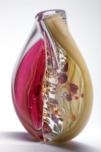 A sculptural blown glass vessel features layers of vibrant pink, shimmering silver leaf, and warm cream colored glass encased in thick clear glass. The twisted form evokes the movement of a woman's hips.
