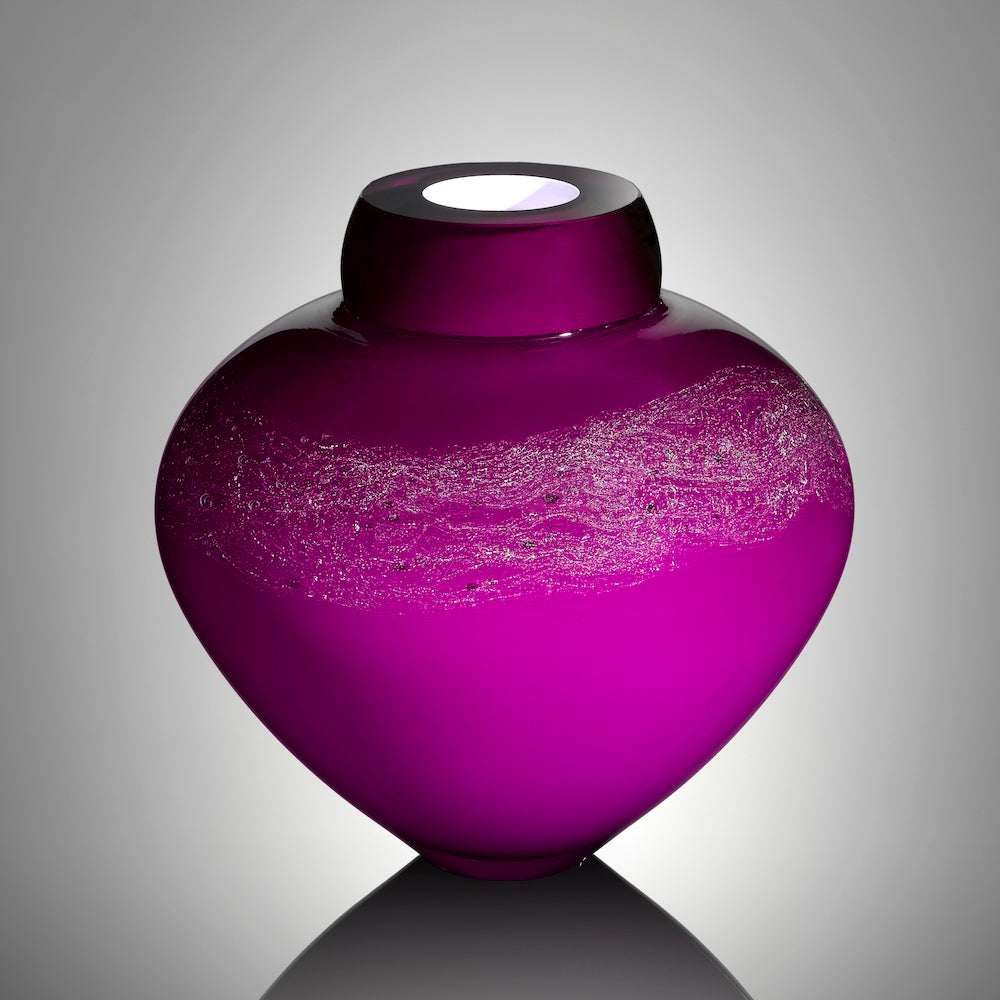 A vibrant purple blown glass vessel adorned with a shimmering band of silver stardust and a bright white interior stands against a gray background.