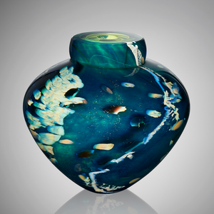 A hand blown glass vessel featuring layers of aquatic blues, greens, and flecks of silver stands against a light gray background.