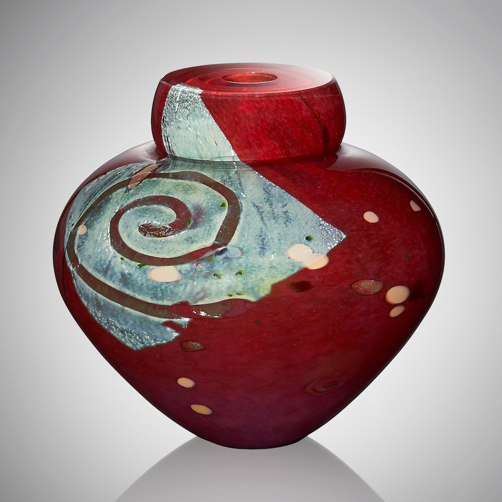 A deep red blown glass vessel features a square of silver leaf decorated with a swirl of clear glass, revealing the red coloration beneath. The surface is adorned with pops of burgundy and apricot glasses.